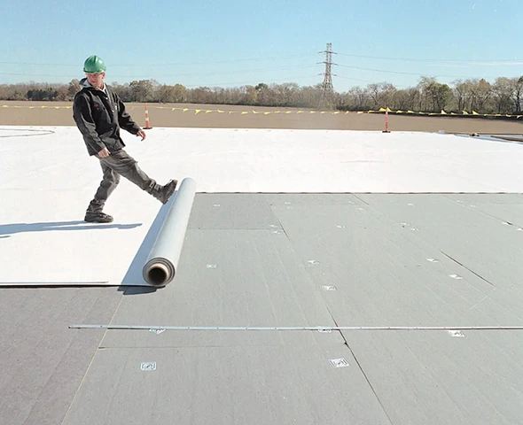 Worker installing PVC roofing on a flat roof, rolling out a white membrane.